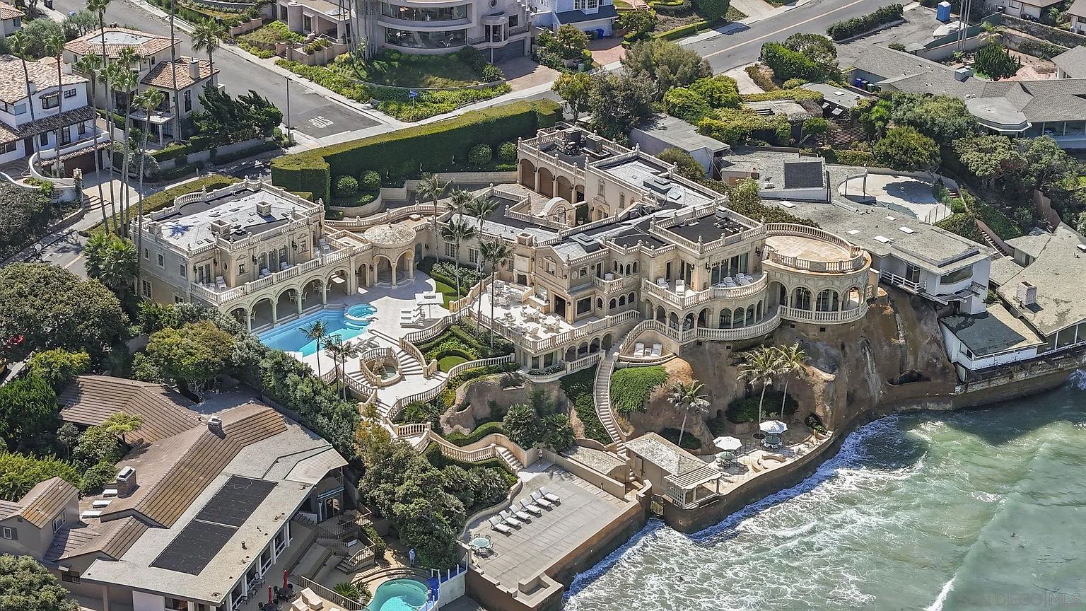 This aerial view showcases a luxurious multi-level estate perched dramatically on a cliffside overlooking the ocean. The property features Mediterranean-inspired architecture with multiple balconies, a swimming pool, meticulously landscaped gardens, and a private beach access area. The overall impression is one of opulence and exclusivity, highlighting the property's unique coastal setting and high-end amenities.