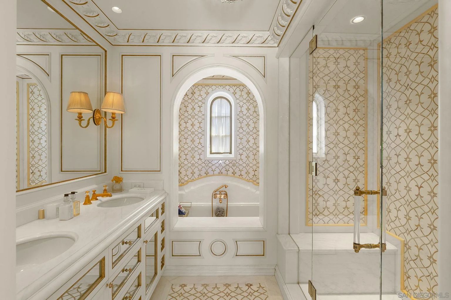 This is a luxurious primary bathroom featuring a double vanity with marble countertops and gold fixtures. An arched alcove houses a soaking tub with a window, and a glass-enclosed shower is visible to the right. The bathroom is decorated in a white and gold color scheme, creating an elegant and opulent atmosphere.