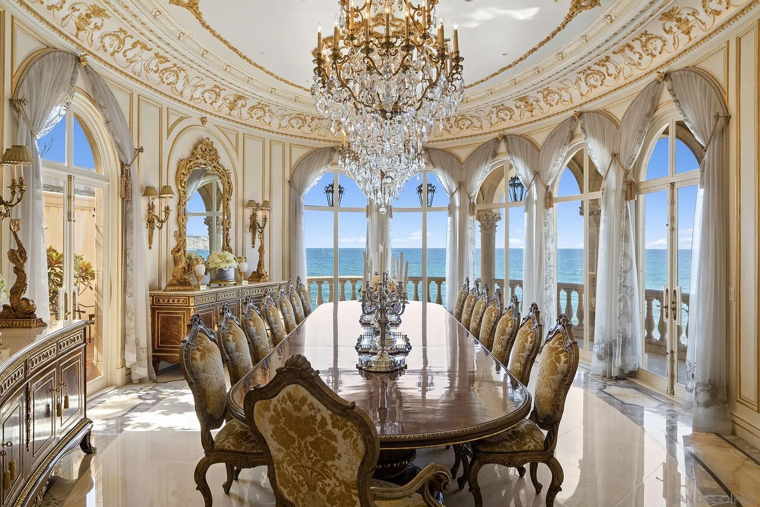 This is a luxurious dining room featuring a long, polished wooden table surrounded by ornate chairs with gold accents. A grand crystal chandelier hangs above the table, and large arched windows offer stunning ocean views. The room is decorated in a classic, opulent style with intricate ceiling details and elegant draperies, creating an atmosphere of grandeur and sophistication.