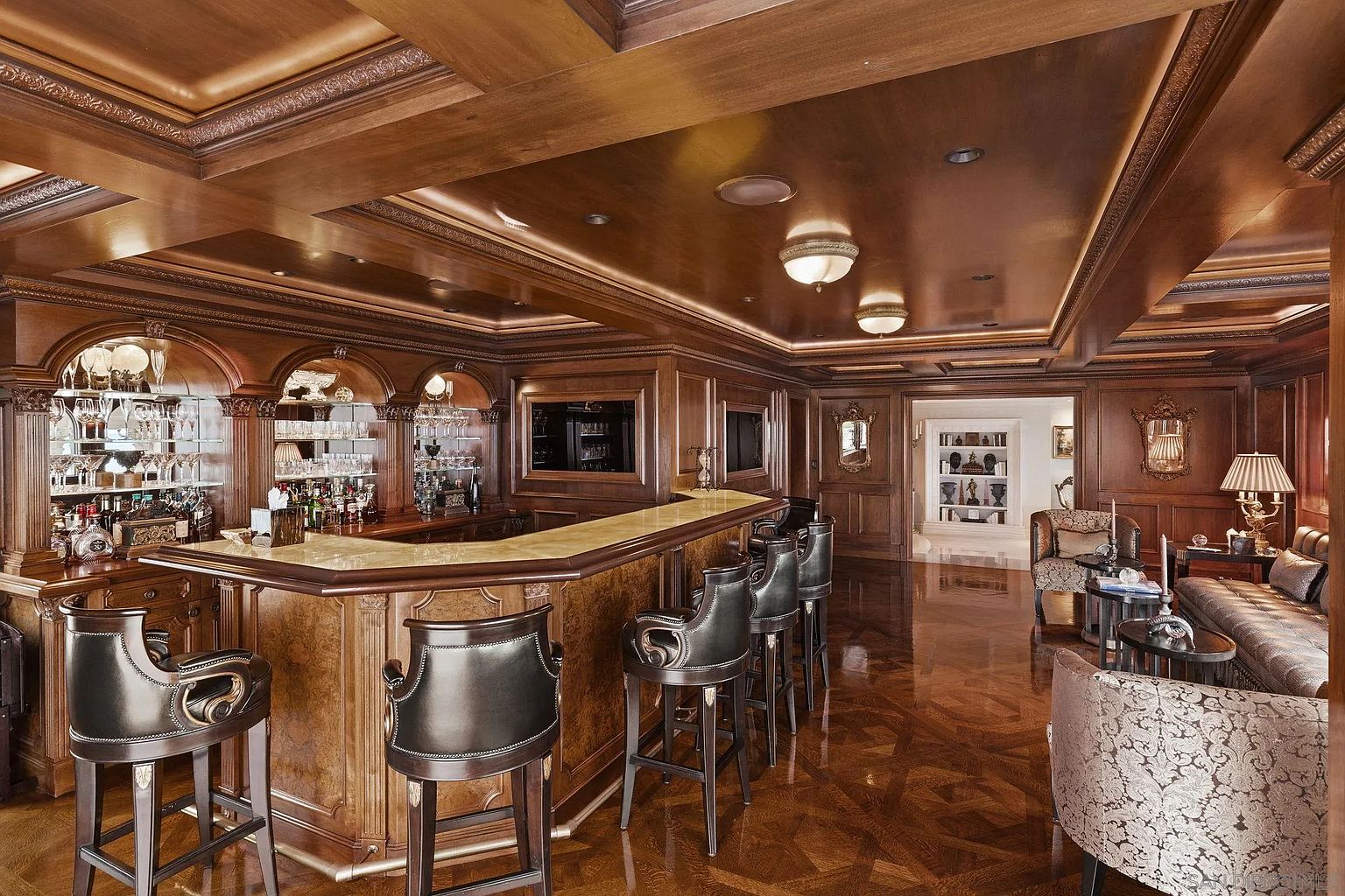 This is an opulent wine cellar featuring rich wood paneling on the walls and ceiling, creating a warm and luxurious ambiance. A well-stocked bar with elegant stools is the focal point, complemented by comfortable seating in the background. The room is illuminated by recessed lighting and decorative fixtures, highlighting the high-end finishes and sophisticated design.