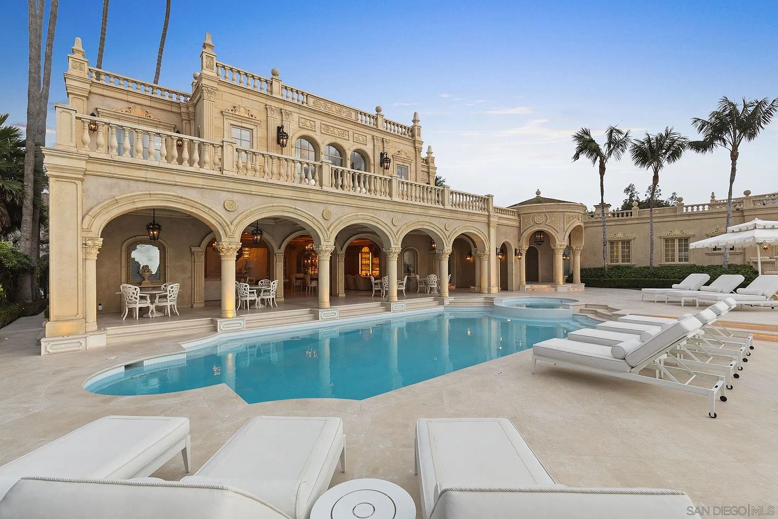 This image showcases a luxurious pool area of an estate, featuring a large, inviting pool with crystal clear water, surrounded by elegant lounge chairs and a meticulously designed patio. The architectural style of the building is reminiscent of a Mediterranean villa, with arched walkways, columns, and a second-story balcony, creating a sophisticated and relaxing outdoor living space. The camera pans across the pool to the building, showing the scale of the property.