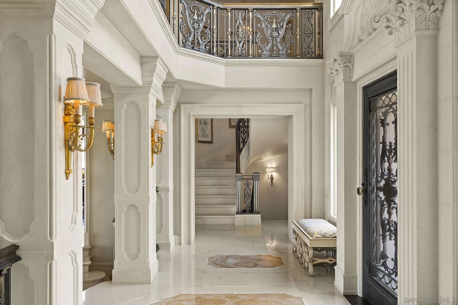 This is an interior shot of a grand hallway featuring marble flooring, ornate columns, and decorative wall sconces. A staircase is visible through an archway, and a wrought-iron balcony overlooks the space, adding to the luxurious and elegant ambiance. The perspective is from the entrance, showcasing the depth and grandeur of the hallway.