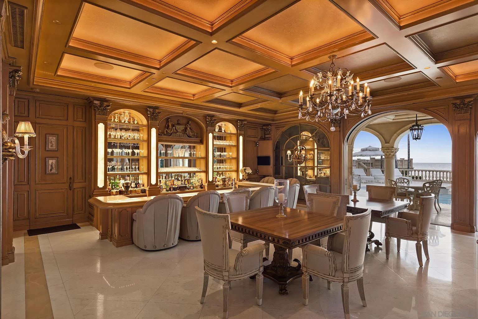 This is an elegant dining room featuring a built-in bar with illuminated shelving, a wooden dining table with upholstered chairs, and a crystal chandelier. The room opens to an outdoor patio with ocean views through an arched doorway. The style is luxurious and traditional, creating a sophisticated ambiance.