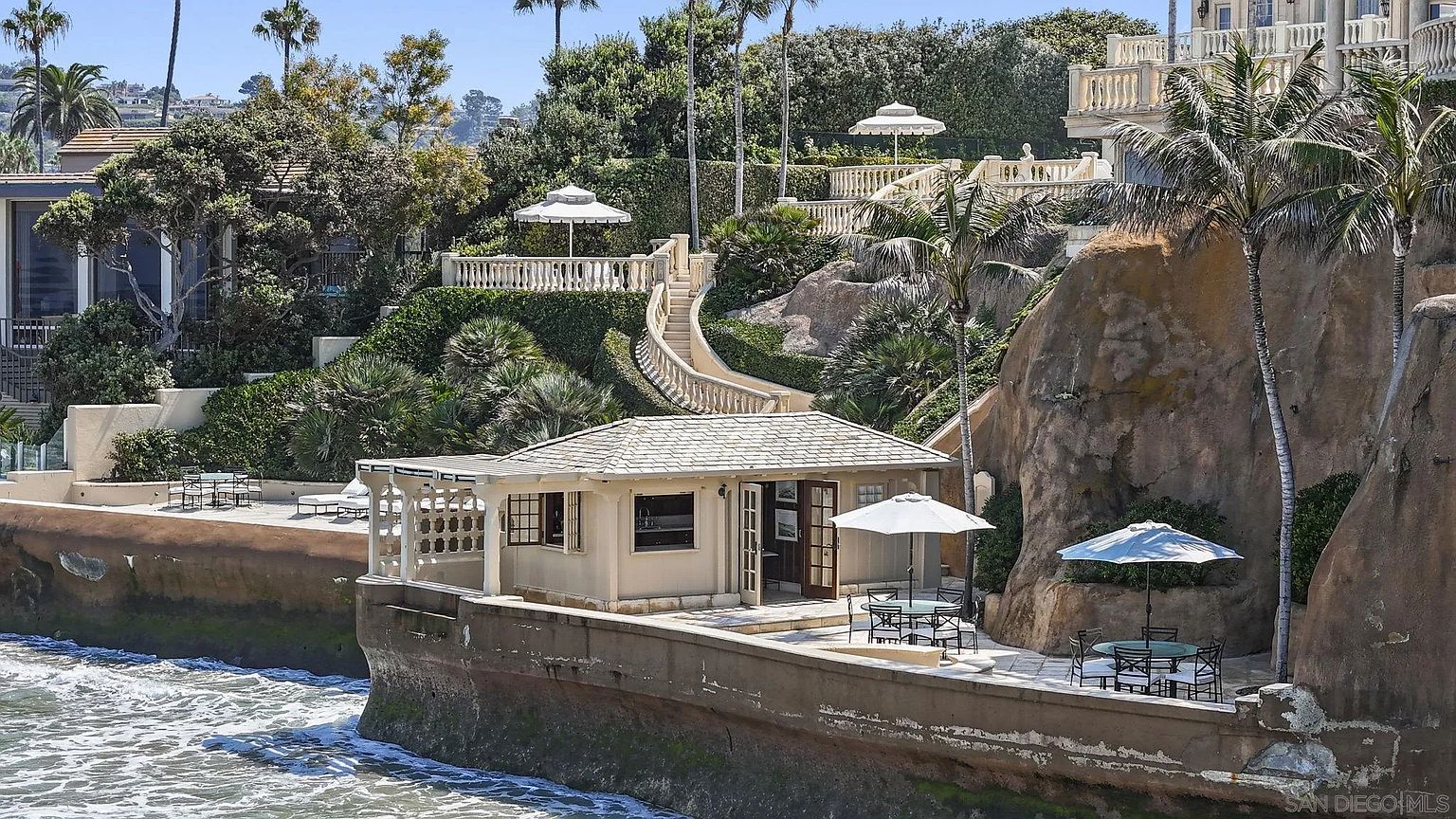 This image showcases an impressive coastal property featuring a charming patio area perched above the ocean. The patio includes outdoor seating arrangements with umbrellas, a small building with an open doorway, and a winding staircase leading up to the main residence. The scene is framed by lush greenery, palm trees, and the rugged coastline, creating a luxurious and serene atmosphere.