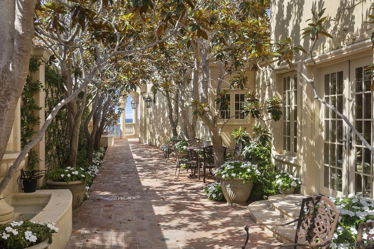 This image showcases a charming outdoor patio area, featuring a brick pathway lined with lush trees and potted plants. The patio includes wrought iron furniture, creating a cozy and inviting space for relaxation or entertaining. In the background, a glimpse of the ocean adds to the serene ambiance, suggesting a luxurious coastal property.