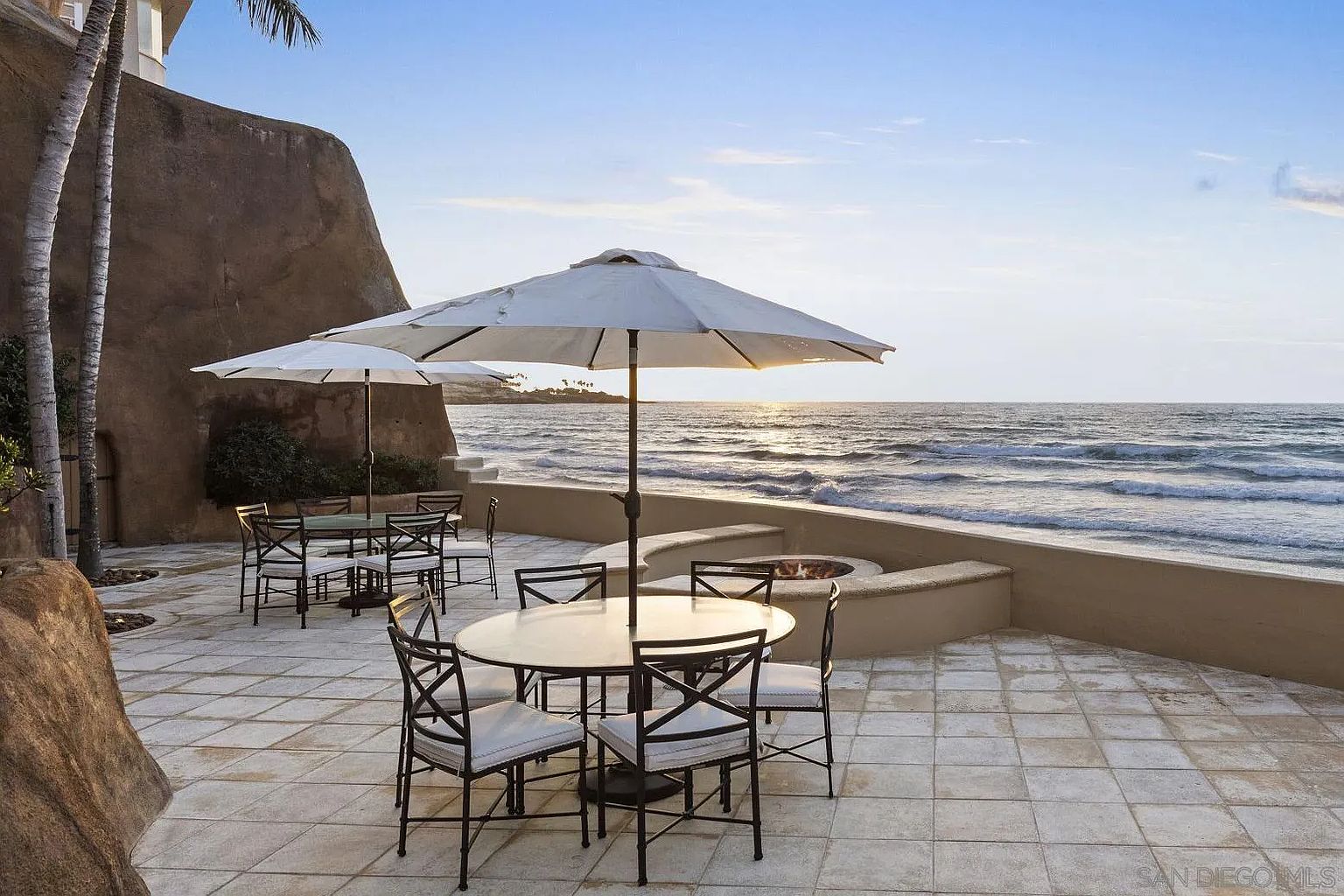 This image showcases a luxurious outdoor patio or deck area with stunning ocean views. The space is furnished with elegant tables and chairs, shaded by large umbrellas, and features a built-in fire pit, creating an inviting atmosphere for relaxation and entertainment. The overall impression is one of upscale coastal living.