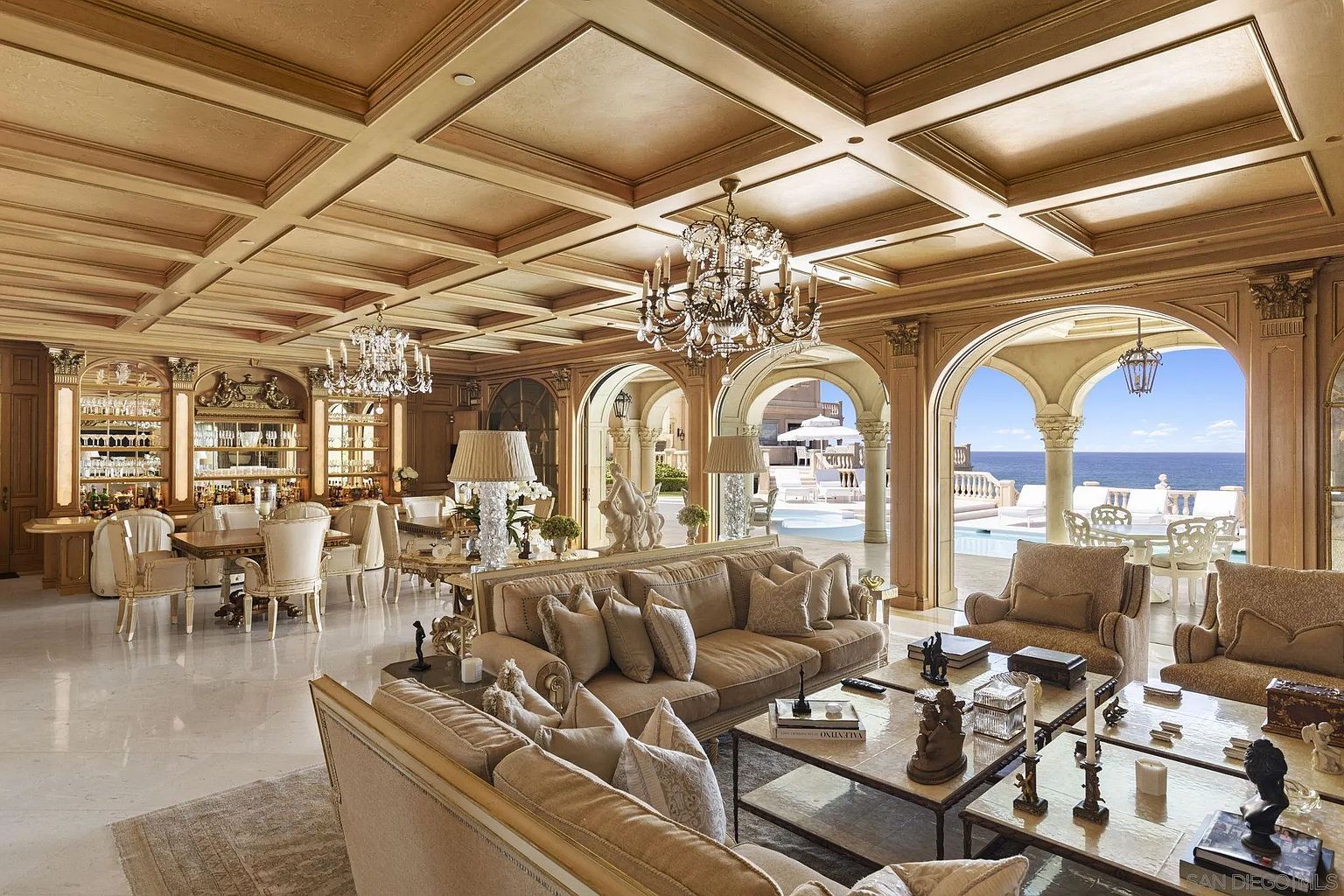 This opulent living room features a coffered ceiling, multiple chandeliers, and arched doorways leading to an outdoor pool area with ocean views. The room is furnished with plush sofas, ornate coffee tables, and decorative accents, creating a luxurious and inviting atmosphere. The color palette is warm and neutral, enhancing the room's elegance.
