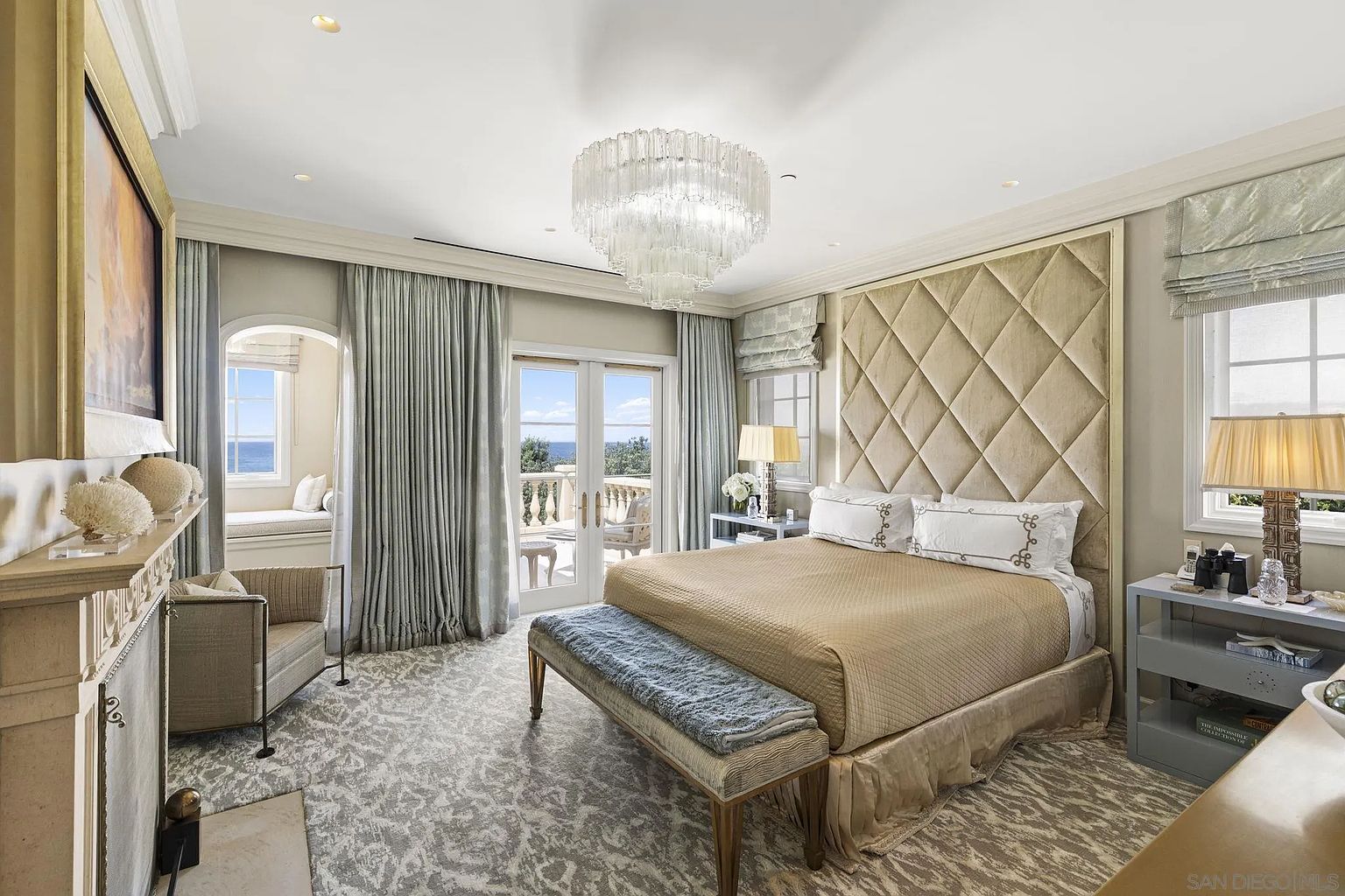 This is a luxurious primary bedroom featuring a large upholstered headboard, a chandelier, and access to a balcony with ocean views. The room is decorated in neutral tones with elegant curtains and a patterned carpet, creating a serene and sophisticated atmosphere. A fireplace and seating area add to the room's comfort and appeal.