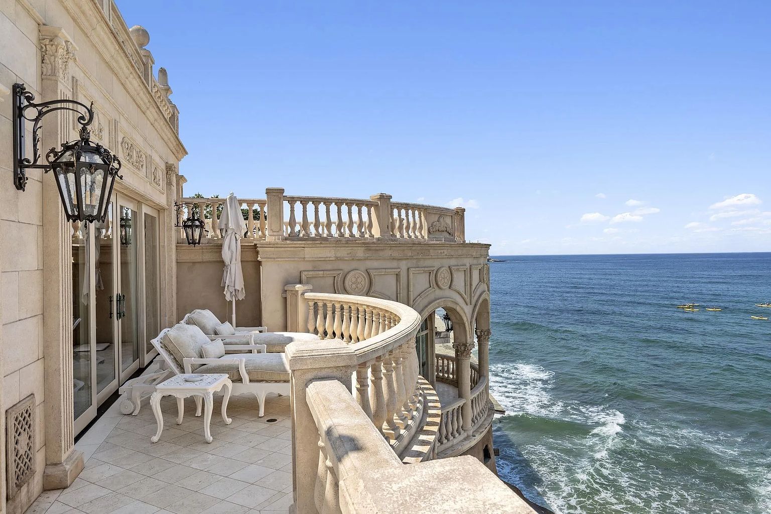 This image showcases a luxurious balcony overlooking the ocean. The balcony features ornate architectural details, including balustrades and decorative moldings. White outdoor furniture, including lounge chairs and a small table, creates an inviting space for relaxation and enjoying the stunning ocean view.