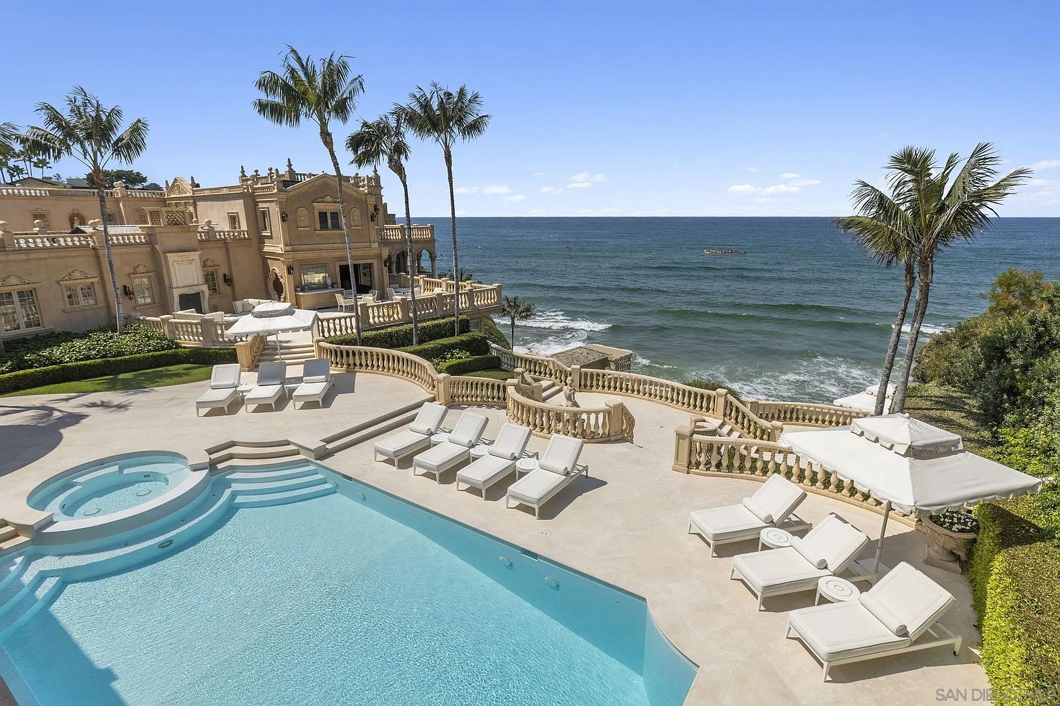 This aerial view showcases a luxurious coastal estate with a stunning pool and spa area. The pool features a unique circular spa and is surrounded by lounge chairs and umbrellas, creating an inviting outdoor space. The property boasts manicured landscaping, a grand Mediterranean-style building, and breathtaking ocean views, emphasizing its high-end appeal.