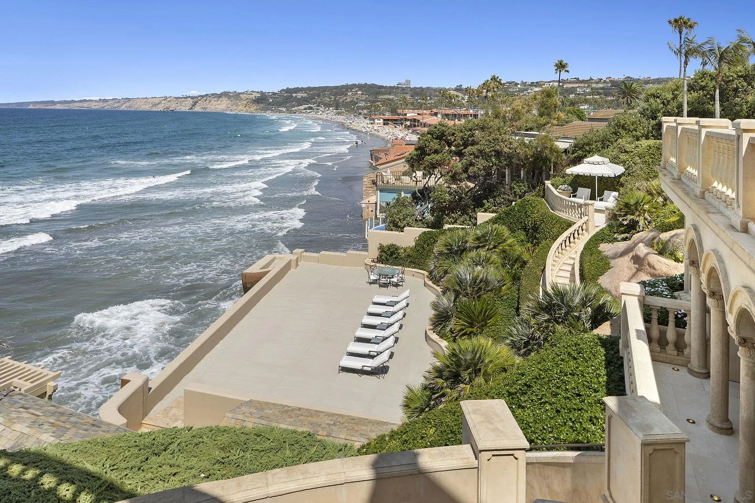 This image showcases a luxurious outdoor patio/deck area with stunning ocean views. The space features multiple lounge chairs, a dining set, and meticulously maintained landscaping. The architectural design includes elegant balustrades and arched walkways, creating a sophisticated and inviting atmosphere perfect for relaxation and entertainment.
