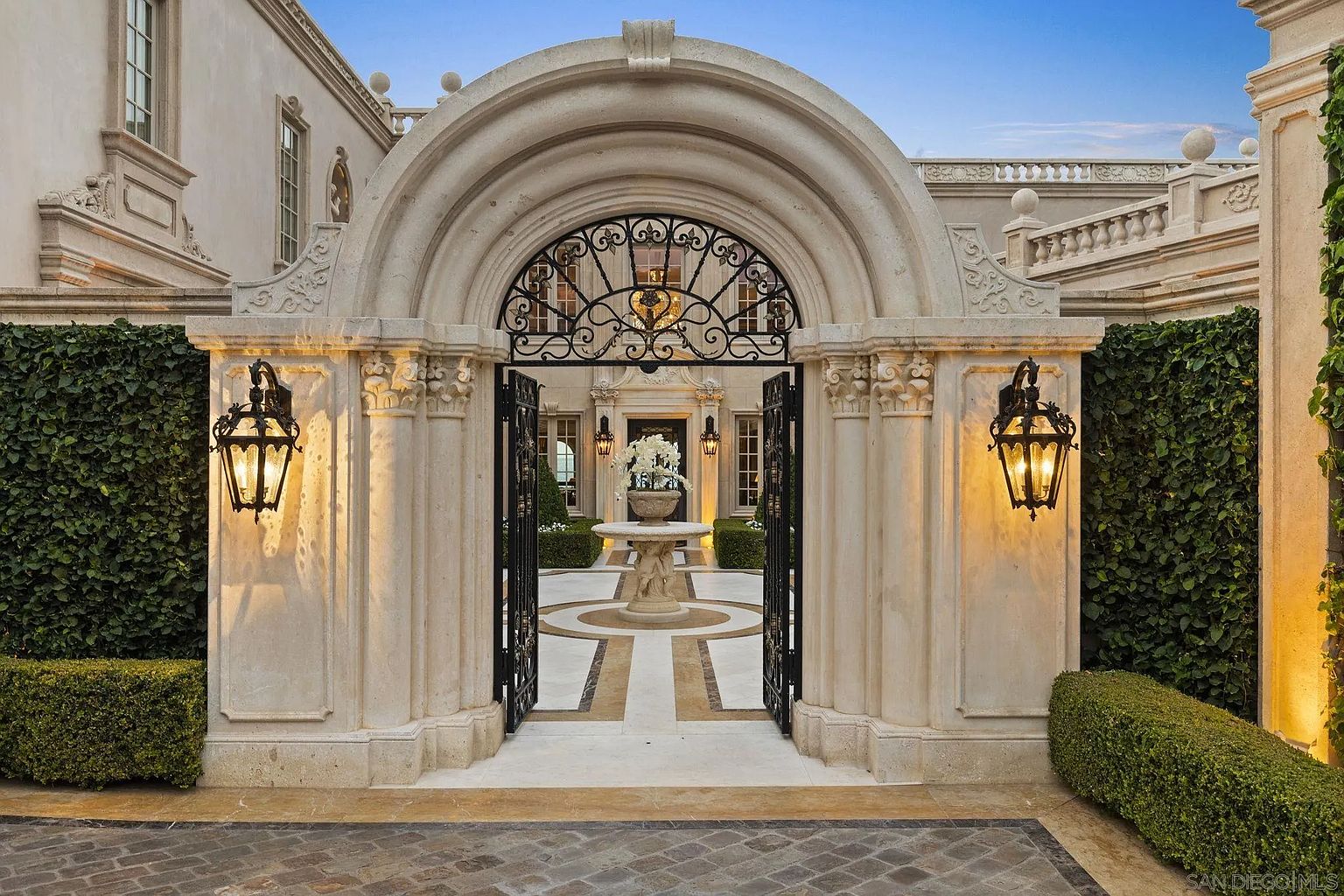 This image showcases a grand entryway to a luxurious estate. An ornate arched gateway with wrought iron gates leads into a courtyard featuring a decorative fountain. The architecture is classical, with stone columns, detailed moldings, and manicured greenery, creating an impression of elegance and exclusivity.