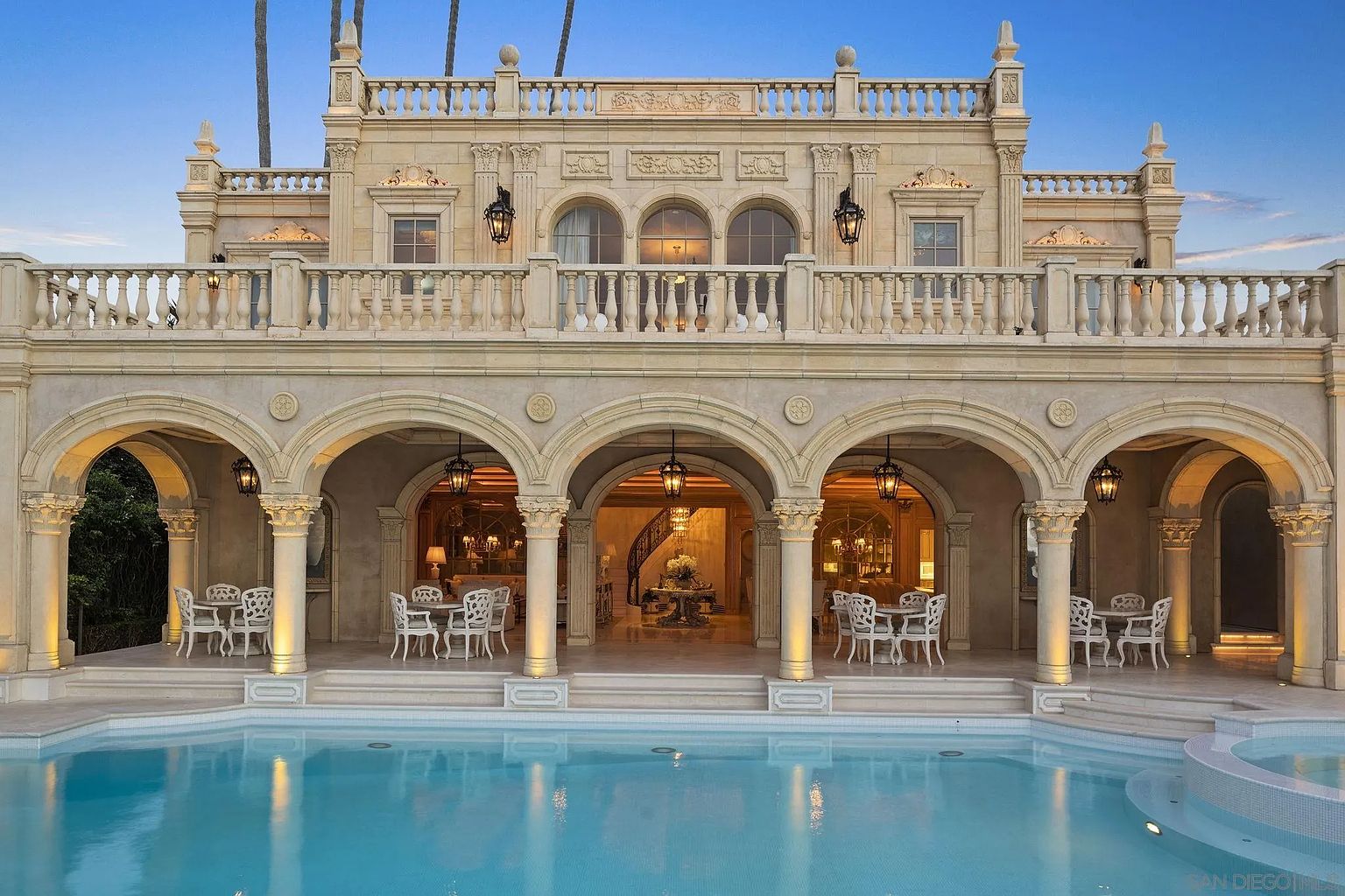 This exterior shot showcases a luxurious mansion's rear facade, featuring a stunning pool and spa area. The architecture boasts elegant arches, columns, and a balcony, creating a grand and inviting atmosphere. The pool's shimmering water reflects the building's design, enhancing the overall opulence and appeal of the property.