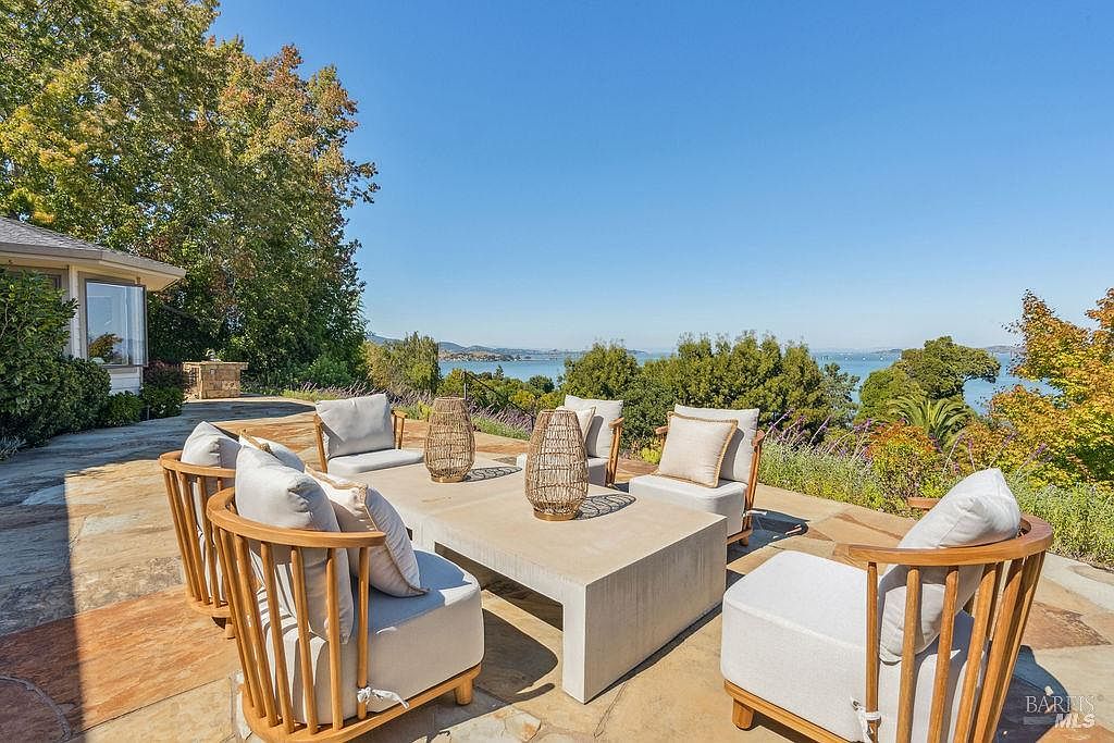 This is an inviting outdoor patio area with comfortable seating arranged around a modern concrete coffee table. The patio is paved with natural stone, and the space offers a beautiful view of the water and surrounding greenery. The scene evokes a sense of relaxation and luxury, perfect for outdoor entertaining.