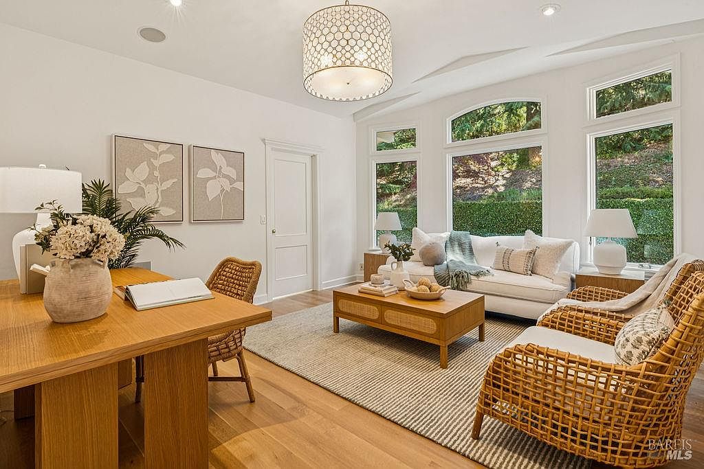This is an inviting living room featuring a neutral color palette with natural wood accents. The room is well-lit with natural light streaming through large windows overlooking lush greenery. The furniture includes a white sofa, woven chairs, and a wooden coffee table, creating a cozy and stylish atmosphere.