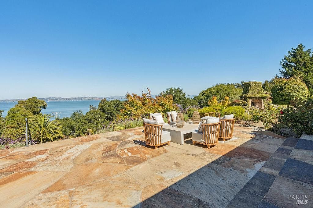 This image showcases a beautifully designed outdoor patio area with a stunning view of the water and surrounding landscape. The patio features stone flooring, comfortable seating arrangements around a modern concrete table, and lush greenery that enhances the natural setting. An outdoor fireplace adds a touch of elegance and warmth to the space, making it an ideal spot for relaxation and entertainment.