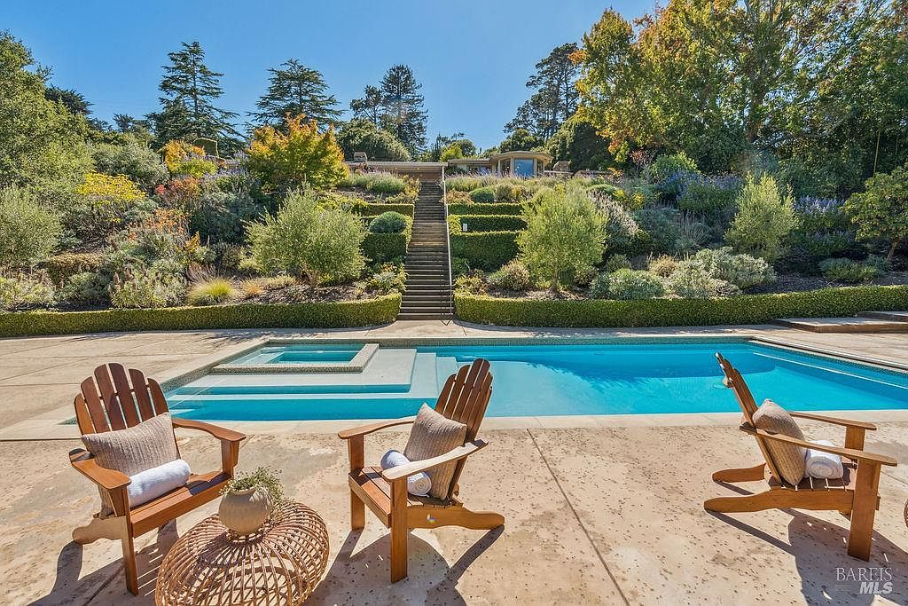 This image showcases a luxurious backyard pool and spa area. Three wooden Adirondack chairs are arranged on the patio, suggesting a relaxing outdoor space. The pool is surrounded by well-manicured landscaping, including tiered gardens and a staircase leading up to the house, creating a serene and private retreat.