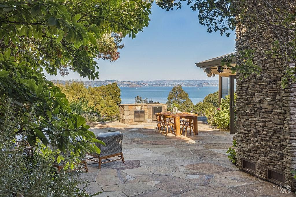 This image showcases a beautiful outdoor patio area with stunning water views. The patio features a stone surface, a built-in outdoor kitchen with a grill, a wooden dining table with chairs, and comfortable lounge seating. A stone wall adds character, while lush greenery frames the scene, creating a serene and inviting atmosphere.