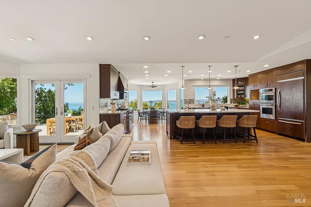 This is an interior shot of an open-concept living room and kitchen area. The living room features a large, comfortable sofa with throw pillows and a blanket, while the kitchen boasts dark wood cabinetry, stainless steel appliances, and a spacious island with bar seating. Large windows and sliding glass doors offer stunning views of the water, creating a bright and airy atmosphere.