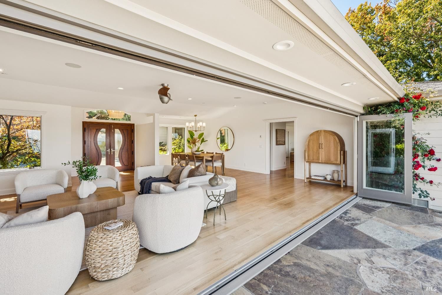 This is a bright and airy living room featuring an open floor plan that seamlessly connects to the outdoors through large sliding glass doors. The room is furnished with comfortable white sofas and chairs, a wooden coffee table, and a woven side table, creating a cozy and inviting atmosphere. The hardwood floors and neutral color palette enhance the sense of spaciousness and modern elegance.