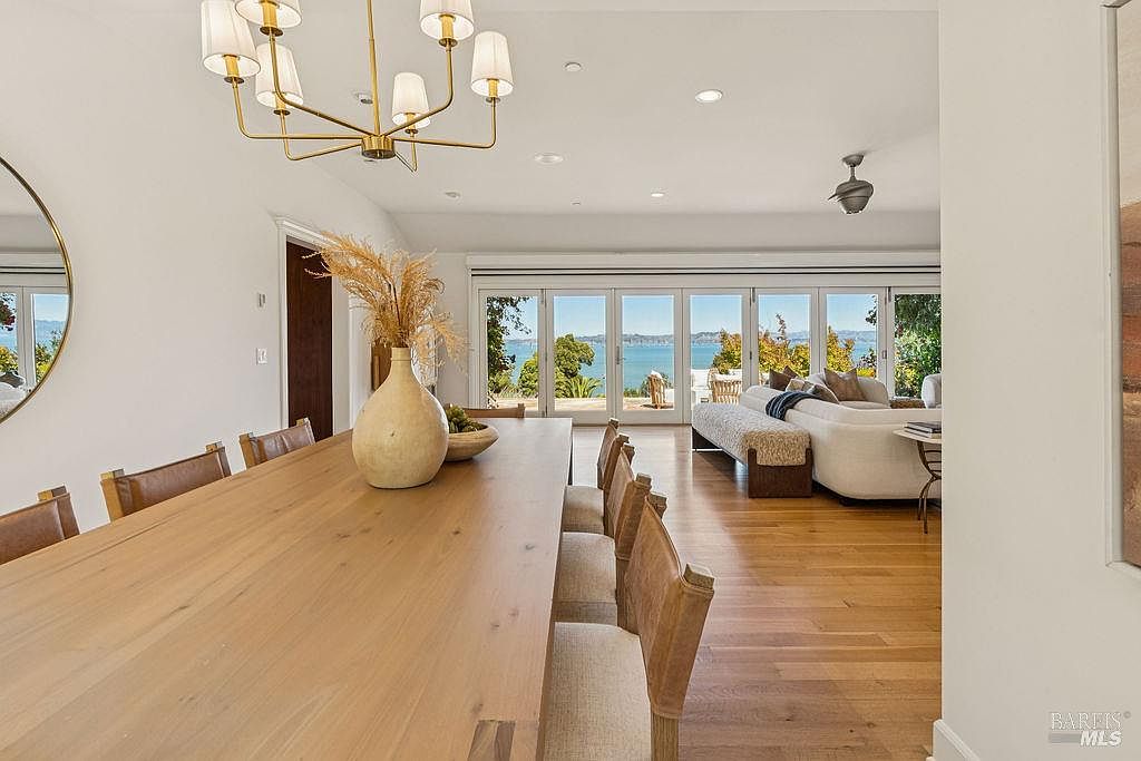 This interior shot showcases a dining room that seamlessly transitions into a living area with a stunning view of the water. The dining room features a long wooden table with upholstered chairs and a modern chandelier, while the living area boasts a comfortable white sofa and large sliding glass doors that open to the outside. The hardwood floors and neutral color palette create a bright and airy atmosphere, perfect for entertaining or relaxing.