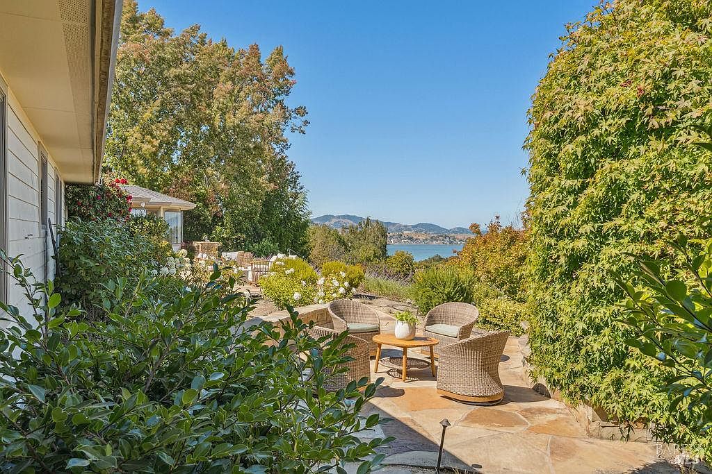 This image showcases a charming patio area with wicker furniture, including chairs and a small table, set on a stone-paved surface. Lush greenery surrounds the patio, providing privacy and a sense of tranquility, while a scenic view of the water and distant hills adds to the appeal. The space is perfect for outdoor relaxation and entertaining.