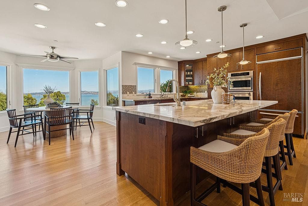 This is a bright and spacious kitchen featuring dark wood cabinetry, a large marble island with seating, and stainless steel appliances. The kitchen also includes a dining area with large windows offering a scenic view of the water. The hardwood floors and recessed lighting add to the luxurious feel of the space.