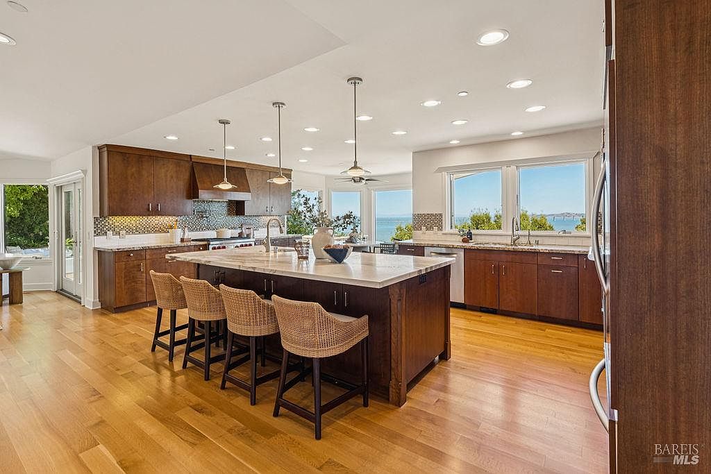 This is a well-lit kitchen featuring dark wood cabinetry and a large center island with a light-colored countertop. The kitchen is equipped with modern appliances and pendant lighting above the island. Large windows offer a view of the water, enhancing the space's appeal.