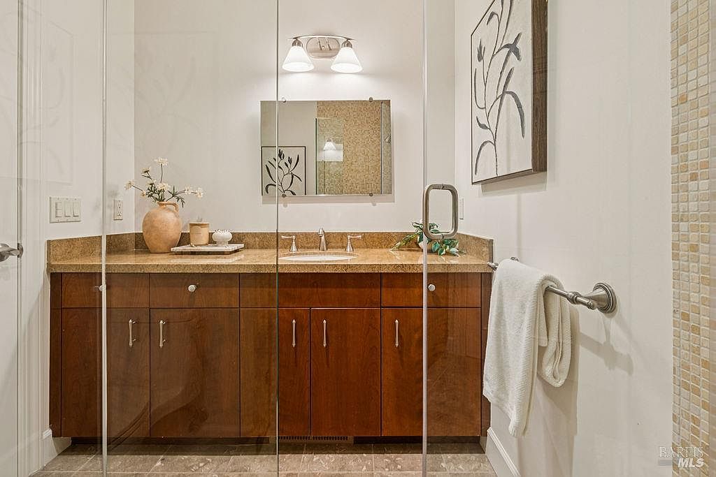 This is a bathroom featuring a vanity with dark wood cabinets and a light-colored countertop. A mirror hangs above the sink, and a decorative painting is on the wall to the right. The shower is enclosed with glass, and a towel hangs on a rack, creating a clean and functional space.