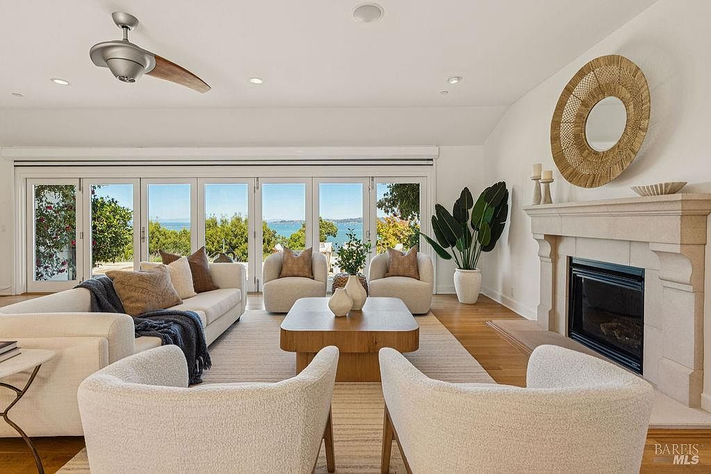 This is an interior shot of a living room featuring a neutral color palette with white walls and light-colored furniture. The room is well-lit with natural light streaming through large glass doors that offer a view of the water. A fireplace with a decorative mirror above it adds a focal point, and the overall style is modern and inviting.