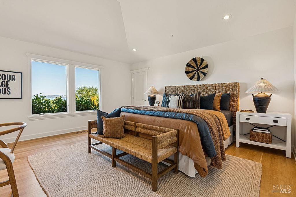 This is a bright and airy primary bedroom featuring a woven headboard and a matching bench at the foot of the bed. The room has a neutral color palette with pops of blue and brown in the bedding and pillows. Natural light floods the space through two large windows, creating a serene and inviting atmosphere.