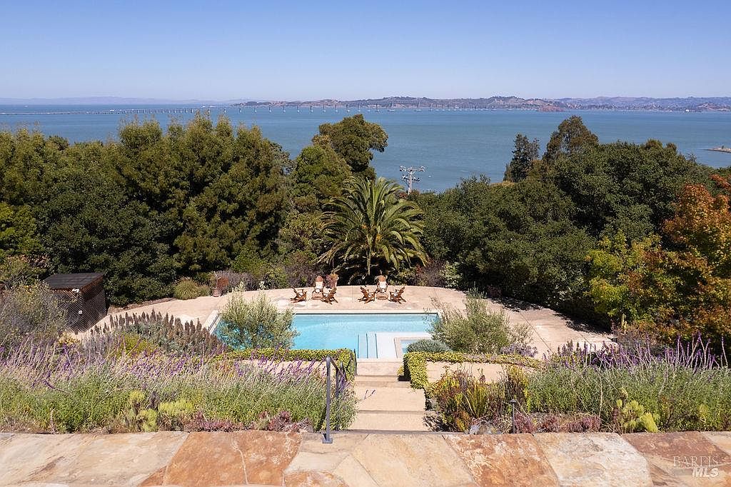 This image showcases a luxurious pool area with a stunning view of the ocean and a bridge in the distance. The pool is surrounded by lush greenery and well-maintained landscaping, creating a serene and private oasis. Several lounge chairs are placed around the pool, inviting relaxation and enjoyment of the picturesque setting.
