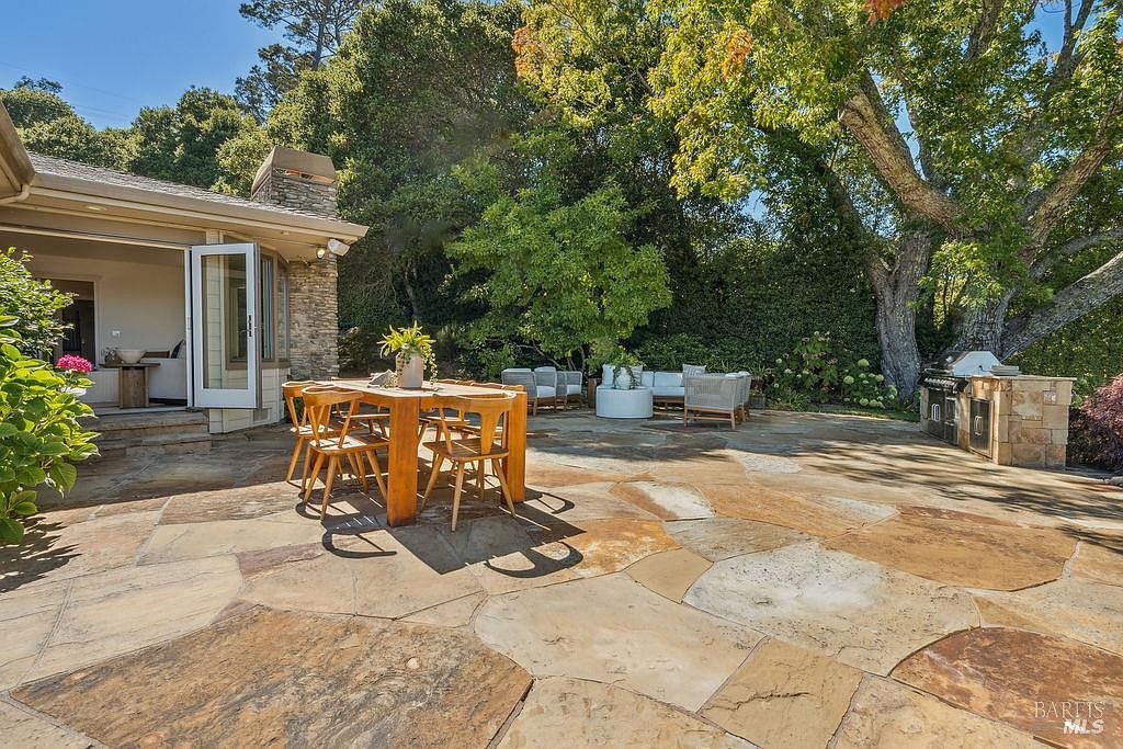 This image showcases a spacious outdoor patio area, perfect for entertaining or relaxing. The patio features a large wooden dining table with chairs, a comfortable seating area with neutral-toned furniture, and a built-in outdoor grill. The stone patio surface and lush greenery surrounding the area create a serene and inviting atmosphere.