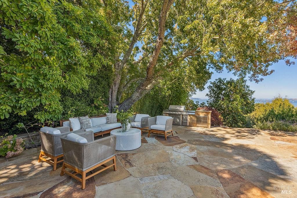 This image showcases a beautifully designed outdoor patio area, featuring comfortable wicker seating arrangements with plush cushions, a modern round coffee table, and a built-in outdoor kitchen with a stainless steel grill. The patio is paved with natural stone, creating a warm and inviting atmosphere, while mature trees provide shade and privacy. The view extends to a distant body of water, enhancing the property's appeal.