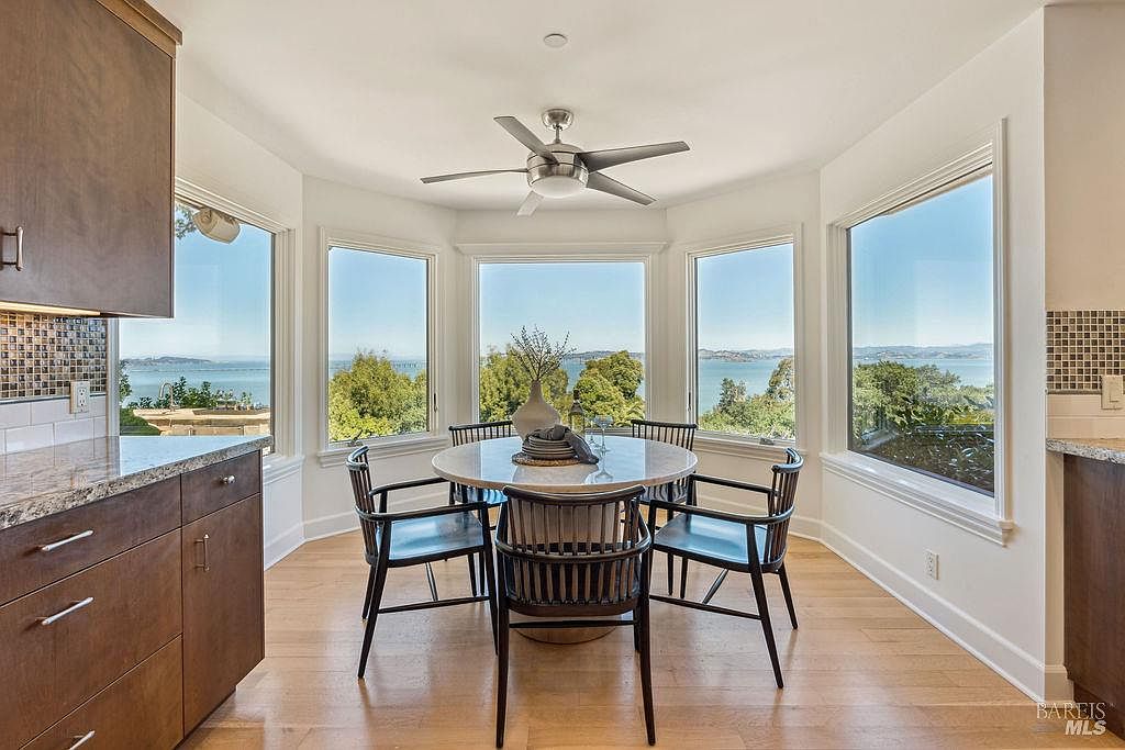 This interior shot showcases a bright dining area with a round table and four chairs, positioned in front of a bay window offering scenic views of the water and landscape. The room features hardwood floors and a modern ceiling fan, creating a cozy and inviting atmosphere. The kitchen area is partially visible on the left.