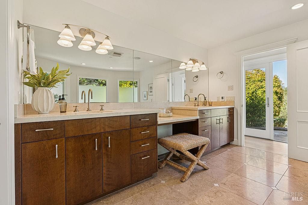 This is a well-lit primary bathroom featuring a double vanity with dark wood cabinetry and light countertops. A large mirror spans the length of the vanity, reflecting natural light from a nearby door leading to the outside. A woven stool is tucked under a makeup area, and the floor is tiled in a neutral tone, creating a clean and inviting space.