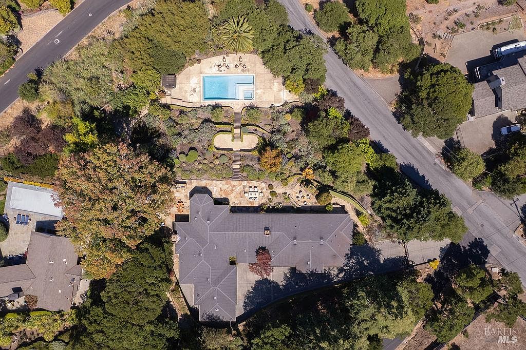 This aerial shot showcases a luxurious property featuring a large house with a dark gray roof, a meticulously landscaped garden with stone pathways, and a stunning pool area. The property is surrounded by lush greenery and mature trees, offering privacy and a serene atmosphere. The image highlights the expansive layout and upscale amenities of the estate.