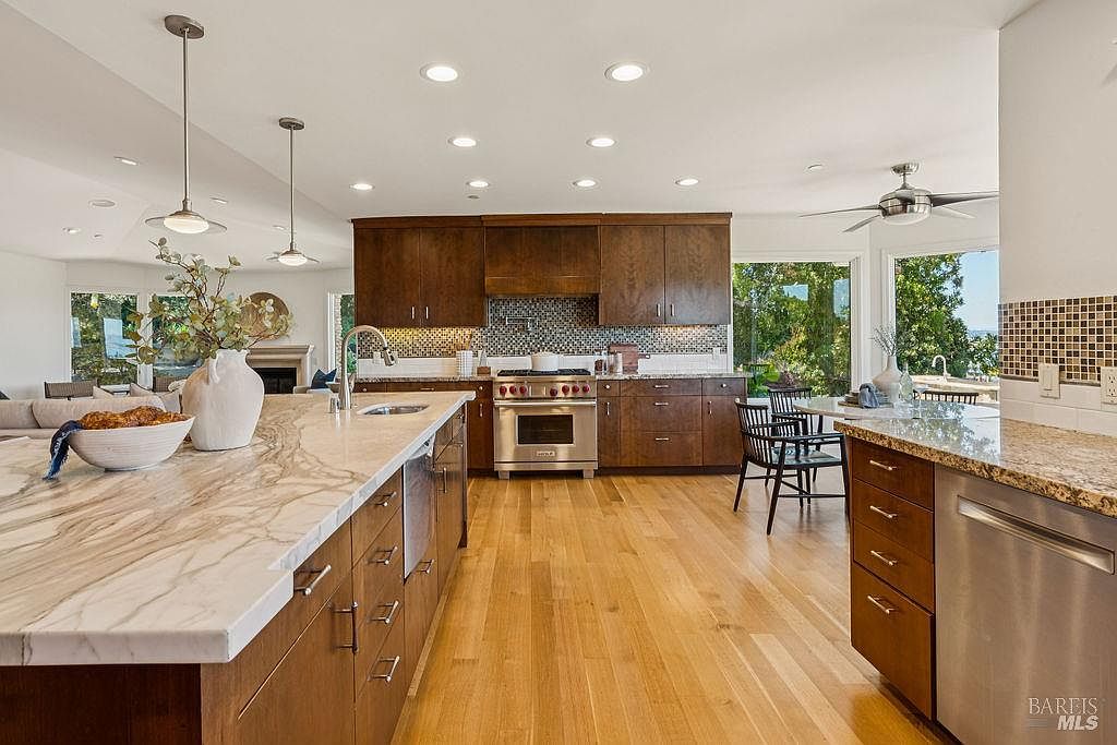 This is a well-lit kitchen featuring dark wood cabinetry, stainless steel appliances, and a large island with a marble countertop. The kitchen has hardwood floors and a mosaic tile backsplash. Large windows provide natural light and views of the outdoors, creating a bright and inviting space.