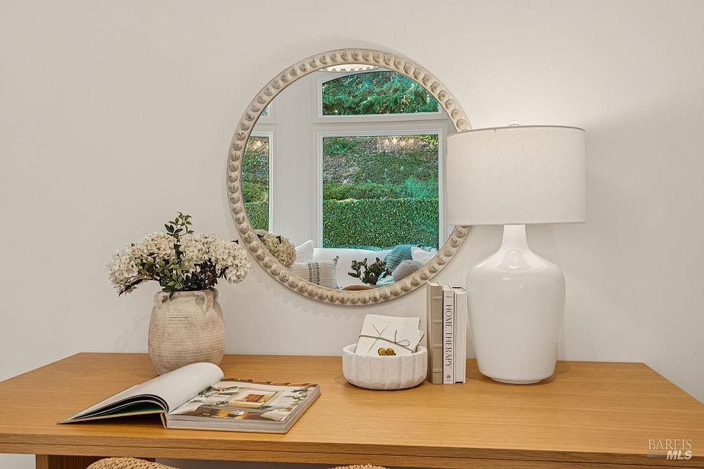 This interior shot showcases a stylish office or study area. A wooden desk is adorned with decorative items such as a vase of flowers, books, and a lamp. A round mirror hangs on the wall, reflecting a view of greenery outside, creating a bright and inviting workspace.