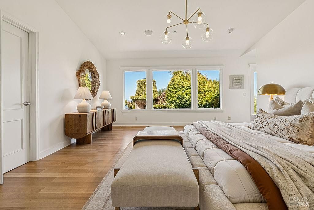 This is a bright and airy primary bedroom featuring hardwood floors, white walls, and large windows offering a view of lush greenery. The room is furnished with a console table with lamps, a decorative mirror, a modern chandelier, and a large bed with neutral-toned bedding and a bench at the foot. The overall impression is one of serene luxury and comfort.