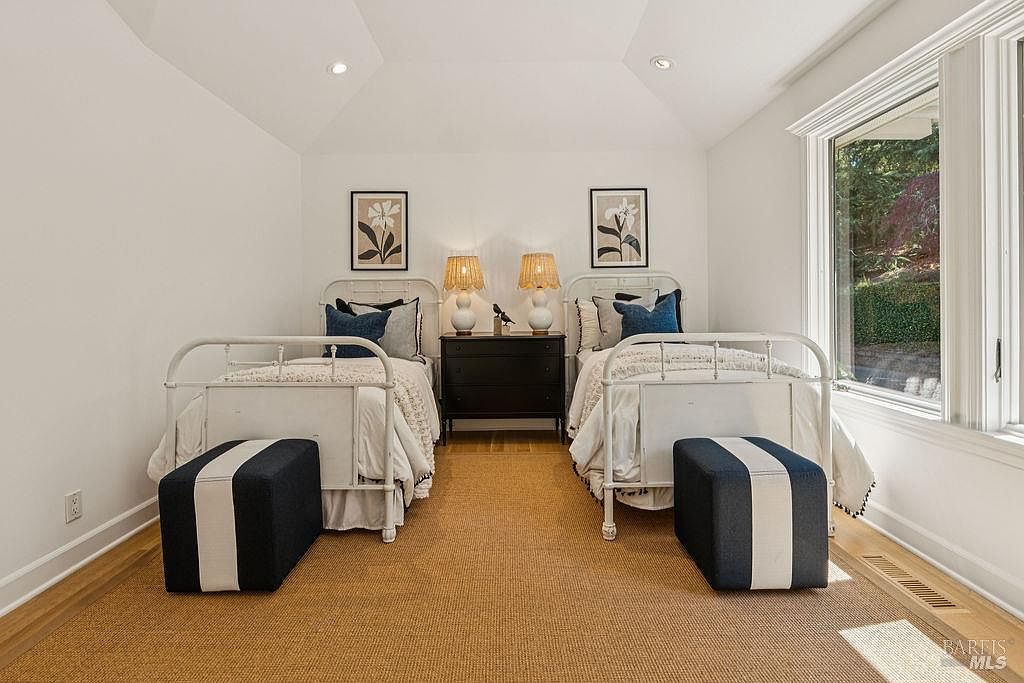 This is an interior shot of a guest bedroom featuring two twin beds with white iron frames, adorned with blue pillows and white bedding. A black nightstand sits between the beds, topped with lamps and decorative objects. The room has a cozy and inviting atmosphere, enhanced by the natural light streaming through the window and the neutral color palette.