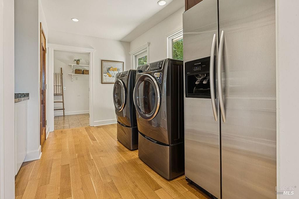 This is an interior shot of a laundry room featuring a modern washer and dryer set in a dark gray finish, positioned next to a stainless steel refrigerator. The room has light hardwood flooring and white walls, with a glimpse into an adjacent room with shelving and a ladder. The overall impression is clean and functional.