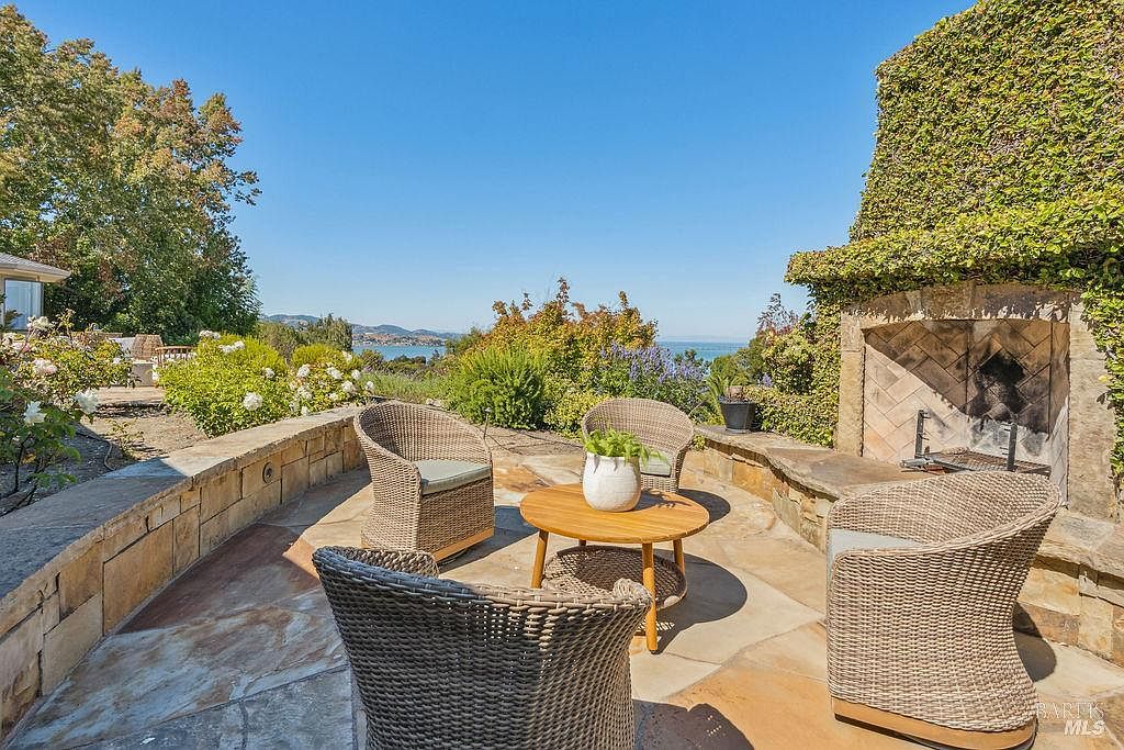 This image showcases a charming outdoor patio area with a stunning view. The patio features comfortable wicker seating arranged around a round wooden table, creating an inviting space for relaxation and conversation. A stone fireplace, partially covered in lush greenery, adds a touch of rustic elegance, while the panoramic vista of the water and distant hills enhances the property's appeal.