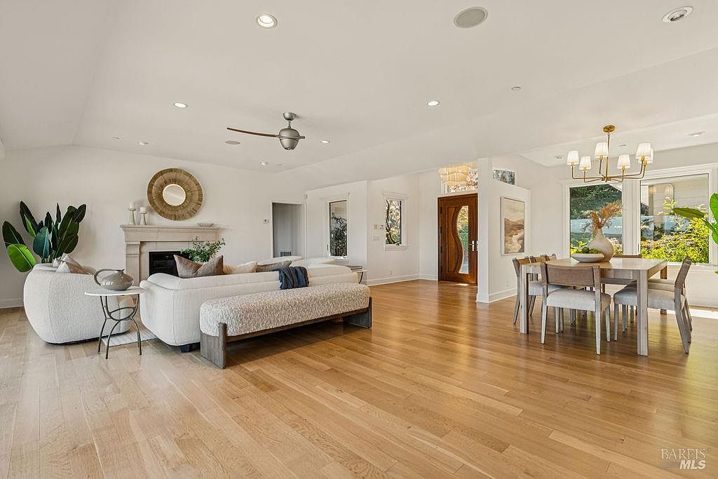 This is an interior shot of a well-lit living and dining area featuring hardwood floors and white walls. The living room area includes a fireplace, a round mirror, a sofa with a bench, and a side table, while the dining area has a wooden table with six chairs and a modern chandelier. The space is open and airy, creating a welcoming and elegant atmosphere.