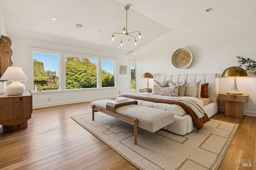 This is a bright and airy primary bedroom featuring a large bed with a tufted headboard, neutral-toned bedding, and a decorative throw. Natural light floods the room through a series of windows, highlighting the hardwood floors and the stylish chandelier. The room is decorated with a woven wall hanging and matching bedside lamps, creating a serene and inviting atmosphere.