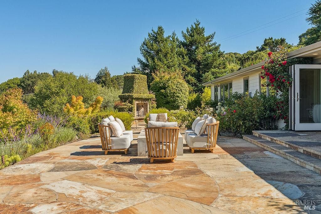 This image showcases a beautifully designed outdoor patio area, featuring comfortable seating arrangements around a central fire pit. The patio is paved with natural stone, blending seamlessly with the surrounding lush greenery and mature trees. A charming outdoor fireplace adds a touch of elegance, while the adjacent building with its open doors suggests easy indoor-outdoor living.