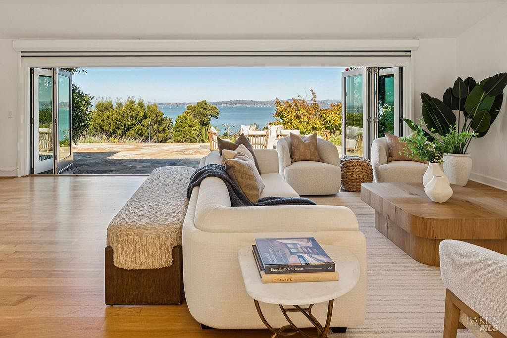 This is an interior shot of a living room featuring a large, open doorway leading to an outdoor patio with a view of the water. The room is furnished with a cream-colored sofa, two armchairs, a wooden coffee table, and a side table with books. The style is modern and luxurious, emphasizing the indoor-outdoor flow and scenic view.