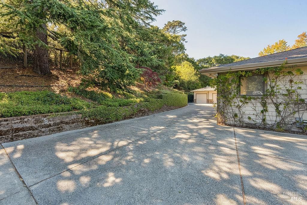This image showcases the side of a house with a concrete driveway leading towards a detached garage. The house is partially covered in climbing vines, adding a touch of natural charm. Lush greenery and trees surround the property, creating a serene and private atmosphere.