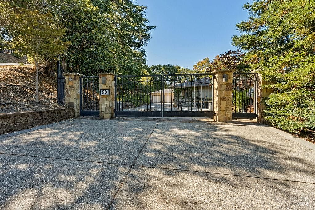 The image showcases an impressive gated entryway to a property. The gate is constructed of dark metal and features stone pillars on either side, with the number '90' prominently displayed. The driveway is paved and leads towards a house partially visible through the gate, surrounded by lush greenery and trees, creating a sense of privacy and exclusivity.