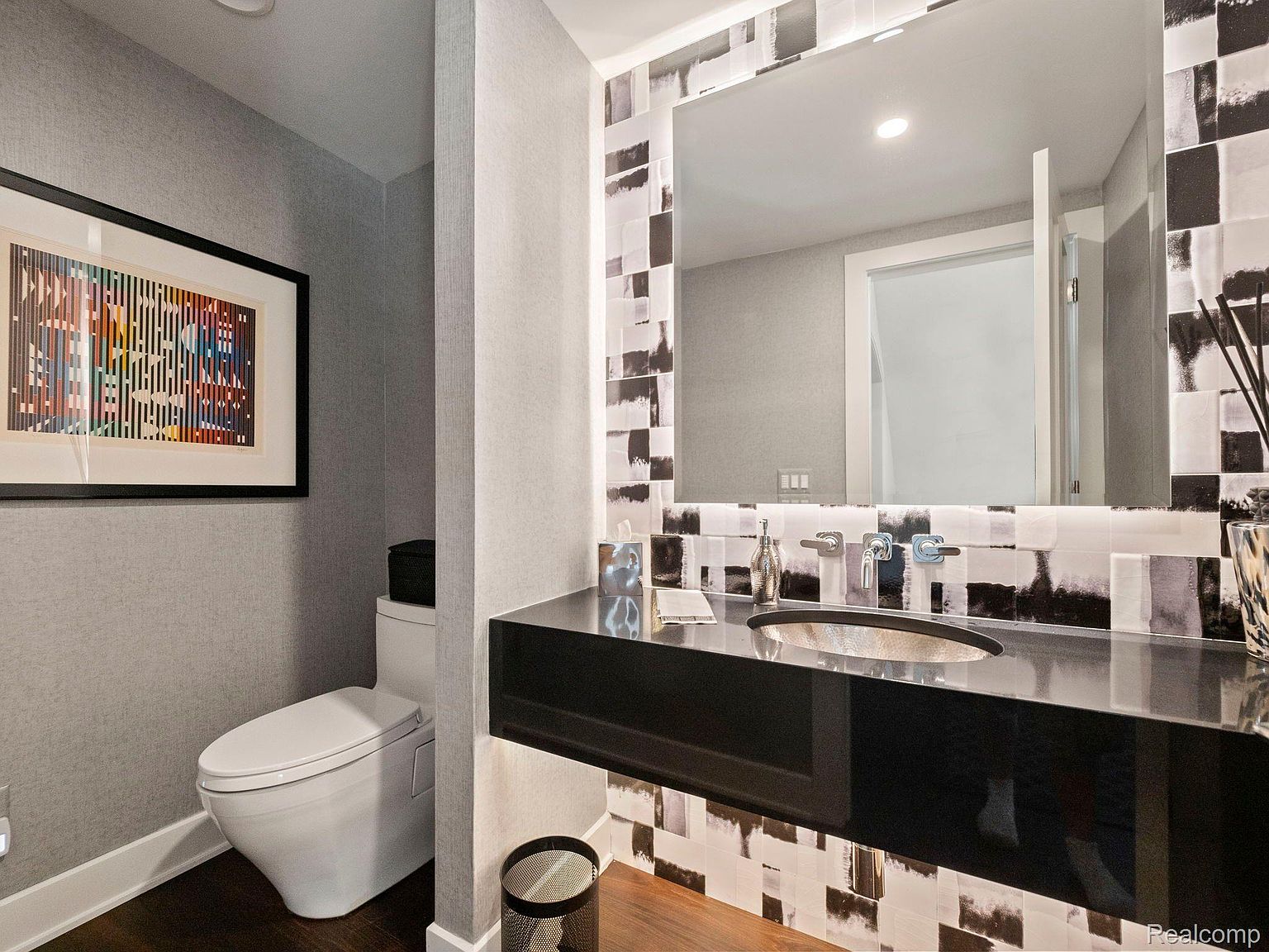 This is a stylish guest bathroom featuring a modern toilet, a sleek black vanity with a vessel sink, and a large mirror framed by black and white patterned tiles. The walls are covered in a textured gray wallpaper, and a framed piece of art hangs above the toilet, adding a touch of sophistication. The dark wood flooring complements the overall contemporary design.